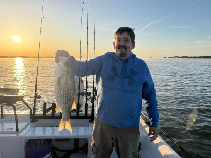 Smiling man in blue hoodie holding a striped bass on a boat during sunset