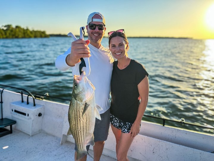 Man and woman smiling on boat holding striped bass at sunset during fishing charter
