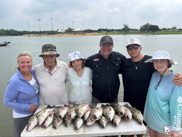 Family of six smiling together on a lake dock with a big catch of fish displayed on a cooler