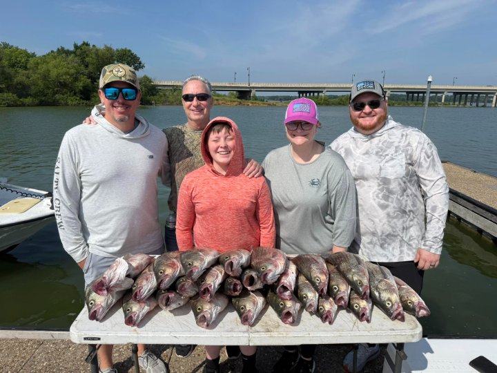 Five happy anglers standing on a dock behind a table full of freshly caught fish