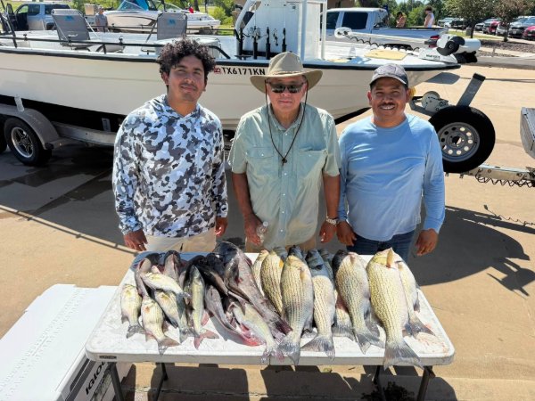Three men standing behind a table displaying a large catch of fish next to a fishing boat