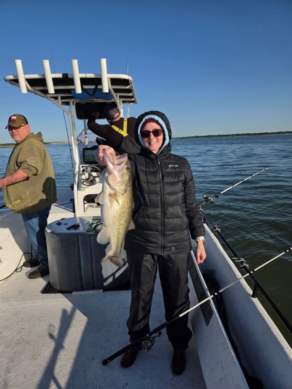 Smiling woman in black jacket holding a large bass on a fishing boat with a man behind her