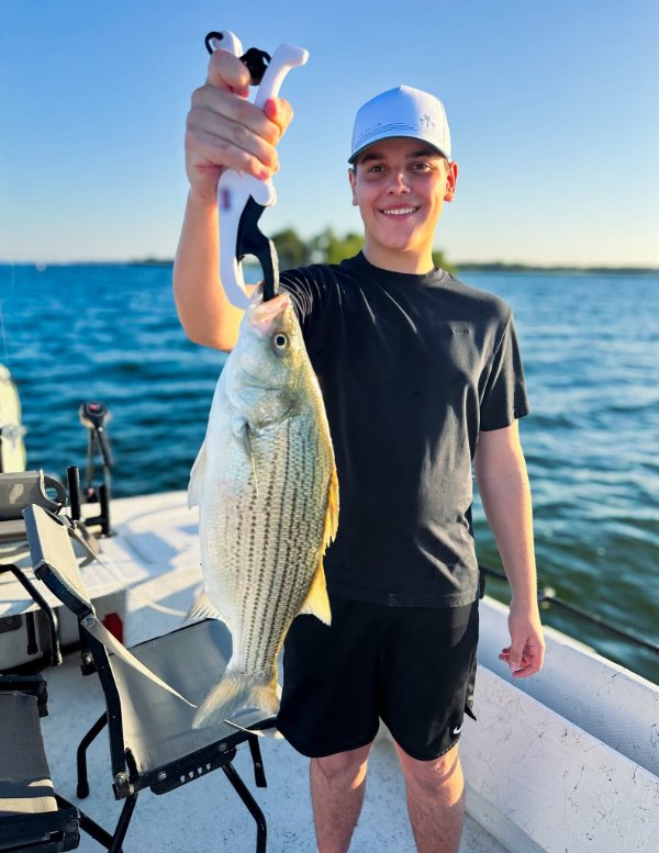 Smiling young man on a boat holding up a freshly caught striped bass with a fish gripper